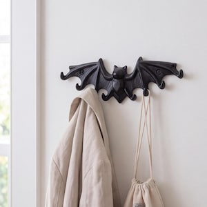 May include: A black, bat-shaped wall hook with a textured finish. The design features outstretched wings that form multiple hooks for hanging items. A beige jacket and a small beige drawstring bag are currently hanging from the bat hooks.