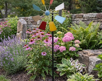 May include: A metal garden wind spinner featuring a central blue hub with eight radiating blades in various colors including yellow, red, teal, tan, and light blue, mounted on a tall, thin black metal stake designed for insertion into the ground.
