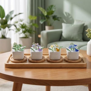 May include: Four small white square planters sit on a rectangular bamboo tray. Each planter contains white filler material, green glass leaves, and a small glass bird in green, purple, yellow, or blue, surrounded by matching glass flowers.