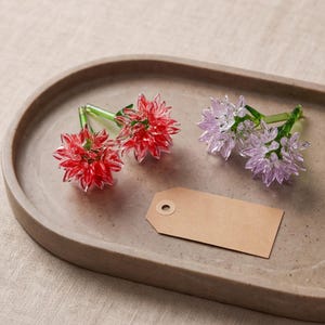 Op de afbeelding: Twee rode glazen bloemfiguren en twee lichtpaarse glazen bloemfiguren, elk met een groene steel, gerangschikt op een gladde, ovale schaal naast een leeg, rechthoekig bruin papieren label met een rond gat aan de bovenkant.