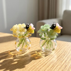 Puede incluir: Dos pequeños jarrones de cristal, cada uno con un ramo de flores y una figura de abeja. Uno tiene flores amarillas, el otro verdes. Las abejas tienen cuerpos a rayas negras y amarillas y alas rosas.