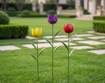 May include: Three metal garden stakes shaped like tulips in yellow, purple, and red. Each flower features a thin green stem with two pointed leaves attached. The stakes are positioned upright in a grassy lawn, displaying vibrant colors and smooth petals.