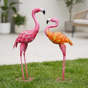 May include: Two metal flamingo sculptures standing on thin legs. The taller flamingo is bright pink with layered feather-like metal plates. The shorter flamingo features a gradient of pink, orange, and yellow feathers. Both have black-tipped beaks.