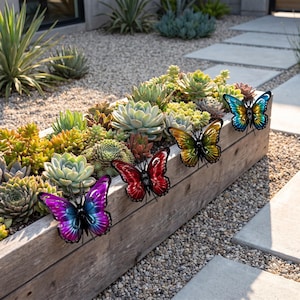 Può includere: Quattro ornamenti a farfalla in metallo fissati sul lato di una fioriera rettangolare in legno riempita con varie piante succulente verdi e verde chiaro. Le farfalle sono colorate in vivaci tonalità viola, rosso, oro e blu con dettagli neri.