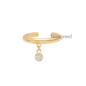 May include: A gold-colored ear cuff with a dangling, round, clear gemstone. The ear cuff has a curved design and the word "Neonjewel" is printed on the side.