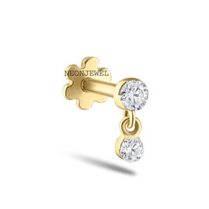 May include: A gold-colored earring with a flower-shaped backing and the text "NEONJEWEL". The earring features a dangling design with two clear, round gemstones. The gemstones are connected by a small, circular link.