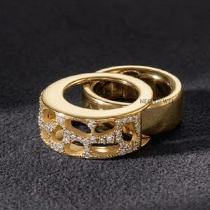 Op de afbeelding: Twee gouden ringen, één met een opengewerkt patroon ingelegd met kleine heldere steentjes en de andere een gladde gepolijste gouden band. De tekst NEONJEWEL is zichtbaar op de zijkant van de gladde band. De ringen liggen tegen elkaar aan.