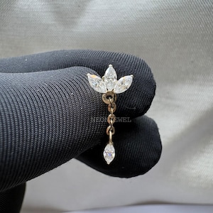 May include: A gold-toned earring featuring a floral design with three marquise-cut white gemstones at the top, connected by a chain to a single dangling gemstone. The earring is set against a dark background.