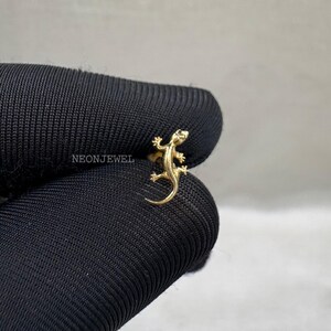Può includere: Un anello a forma di lucertola color oro. L'anello presenta un design dettagliato di lucertola con zampe, coda e testa visibili. L'anello è posto su uno sfondo nero. Il testo "NEONJEWEL" è visibile.