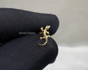 May include: A gold-colored lizard ring. The ring features a detailed lizard design with visible legs, tail, and head. The ring is set against a black background. The text "NEONJEWEL" is visible on the background.