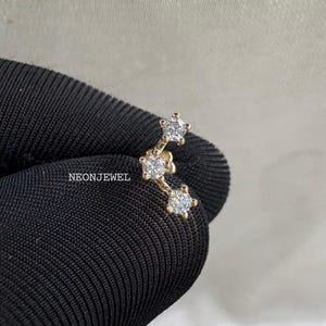Pode incluir: Um brinco trepador dourado com três pedras preciosas claras e brilhantes. O brinco é curvo e tem o texto "NEONJEWEL" em um fundo de tecido preto.