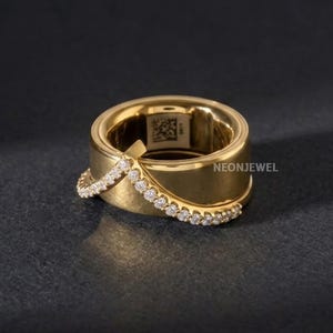 May include: A gold-toned ring featuring a wide, smooth band with a V-shaped overlay encrusted with small, clear, round-cut stones. The interior of the band displays a small square QR code and the text NEONJEWEL printed on the side.