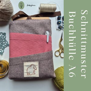 Può includere: Una copertina per libri in tessuto fatta a mano, grigia e rosa, con una piccola toppa a forma di coniglio. La copertina ha una tasca e un portapenne. L'immagine mostra anche forniture per cucire come filo, forbici e bottoni. Il testo laterale recita "Schnittmuster Buchhülle A6".