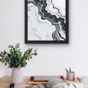 May include: Abstract black and white art print in a black frame. The artwork features a flowing, marbled design. The print is displayed above a wooden desk with a laptop, open book, and a vase of greenery, suggesting a home office or creative space.