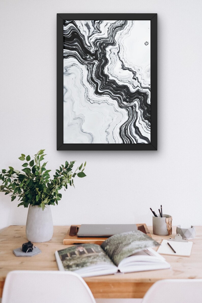 May include: Abstract black and white art print in a black frame. The artwork features a flowing, marbled design. The print is displayed above a wooden desk with a laptop, open book, and a vase of greenery, suggesting a home office or creative space.