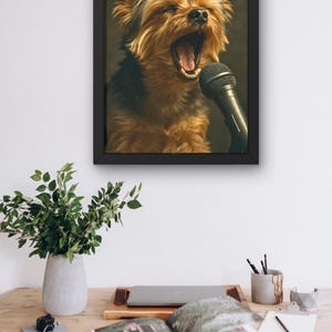 May include: A framed print featuring a Yorkshire Terrier with its mouth open, appearing to sing into a microphone. The dog has brown and black fur. The black frame is hanging on a white wall above a wooden desk with a plant, laptop, and open book.
