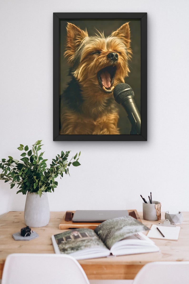 May include: A framed print featuring a Yorkshire Terrier with its mouth open, appearing to sing into a microphone. The dog has brown and black fur. The black frame is hanging on a white wall above a wooden desk with a plant, laptop, and open book.