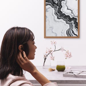 May include: A framed abstract art print with black and white swirling patterns hangs on a white wall. A glass vase with pink flowers, a green drink, and a matcha whisk sit on a gray table. A person is in the foreground.
