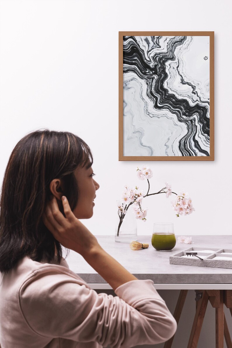 May include: A framed abstract art print with black and white swirling patterns hangs on a white wall. A glass vase with pink flowers, a green drink, and a matcha whisk sit on a gray table. A person is in the foreground.