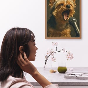 May include: A framed print depicts a Yorkshire Terrier singing into a microphone. The dog has brown and black fur and is mid-yawn. A woman is seen in profile, looking at the print. A glass of green liquid and a vase of flowers are on the table.