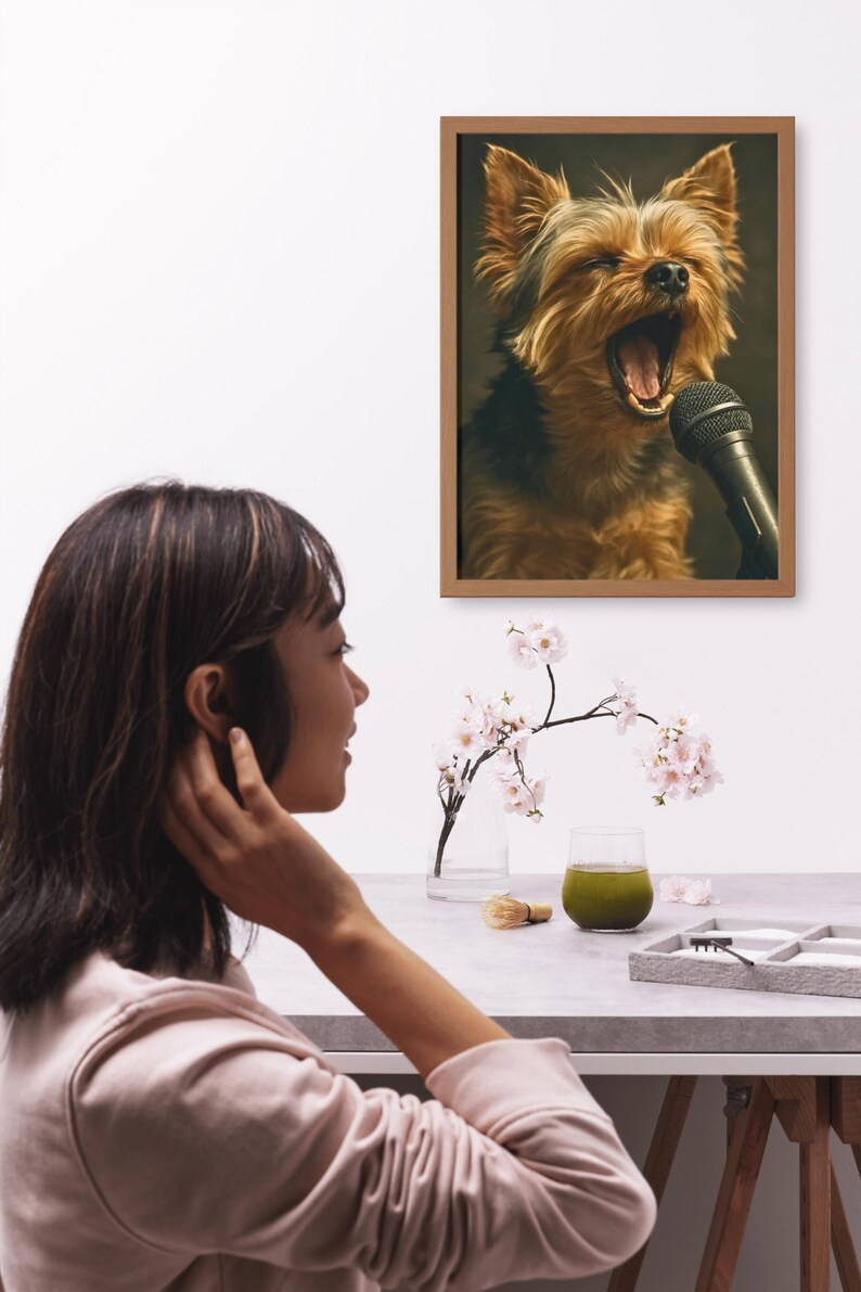 May include: A framed print depicts a Yorkshire Terrier singing into a microphone. The dog has brown and black fur and is mid-yawn. A woman is seen in profile, looking at the print. A glass of green liquid and a vase of flowers are on the table.