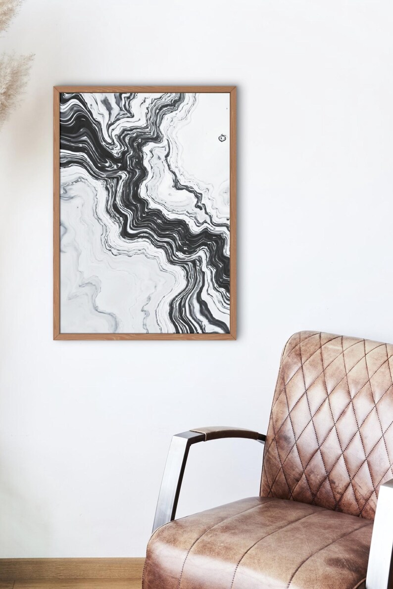 May include: Abstract art print in a wooden frame. The artwork features a flowing, marbled design in shades of black and white. The print is displayed on a white wall, with a brown leather armchair in the foreground.