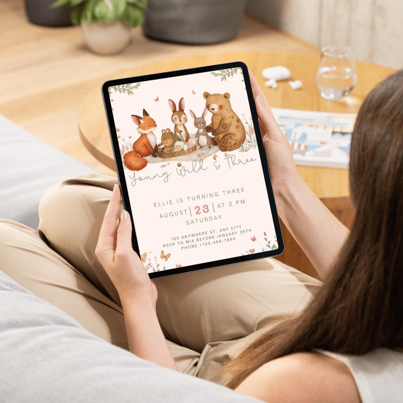 May include: A tablet displays a birthday invitation with woodland animal illustrations. The invitation reads "Young Wild & Three" and includes details like the date, time, and RSVP information. The design features a fox, bear, and rabbits.