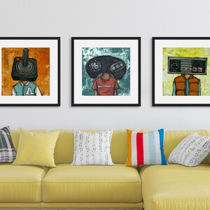 May include: Three framed illustrations of people with video game controllers for heads. The first illustration is a person with a joystick for a head, wearing a blue and orange letterman jacket. The second illustration is a person with a Sega Genesis controller for a head, wearing a red shirt. The third illustration is a person with a Nintendo Entertainment System controller for a head, wearing a brown vest and a blue shirt.
