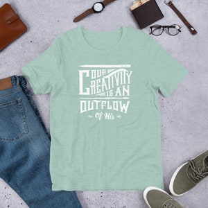 Op de afbeelding: Een lichtgroen T-shirt met een wit grafisch ontwerp met de tekst "Onze creativiteit is een uitstroom van Hem".
