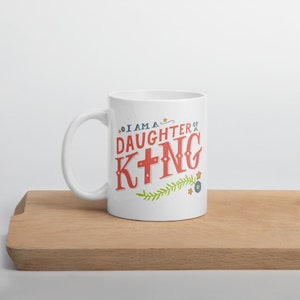 May include: White ceramic mug with a red and blue design that says "I am a daughter of a King" with a cross in the word King. The design also includes green leaves and blue flowers.