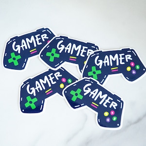 May include: A collection of navy blue controller stickers, each featuring the word "GAMER" in white. The controllers have green directional pads, colourful buttons, and a white outline. The stickers are arranged on a white surface.