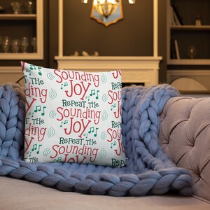 May include: A white throw pillow with a repeating pattern of the words "Sounding Joy" in red and green text with musical notes. The pillow is on a light blue couch with a white throw blanket.