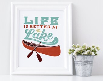 May include: White framed print with the text "Life is better at the Lake" in a vintage font. A red canoe with two oars is pictured below the text.
