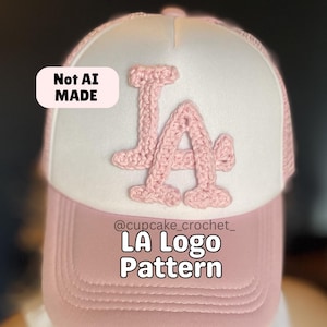 May include: A pink and white trucker hat with a crocheted "LA" logo. The hat features a pink brim and a white front panel with a pink crocheted "LA" logo. The text "LA Logo Pattern" is displayed on the hat.