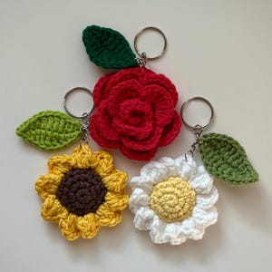 May include: Crocheted flower keychains in red rose, yellow sunflower, and white daisy designs. Each flower has a green leaf and a silver keyring. The flowers are handmade and have a textured appearance.