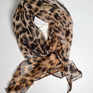 Può includere: Sciarpa trasparente con stampa leopardata nei toni del marrone e del nero. La sciarpa è drappeggiata, con un'etichetta attaccata. L'etichetta recita "Silk Pillow".