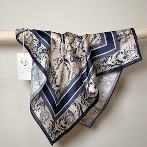 May include: A square silk scarf with a cream background and a navy blue border and design. The scarf features a repeating pattern of suns, plants, and other decorative elements. The tag reads "All the Silk".