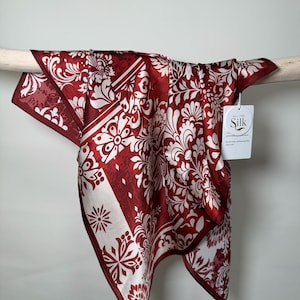 Può includere: Una sciarpa quadrata di seta in una tonalità rosso intenso, ornata da un motivo floreale e a volute bianco. La sciarpa è drappeggiata su un'asta di legno, mostrando il suo design intricato. Un'etichetta con la scritta "All the Silk" è attaccata.