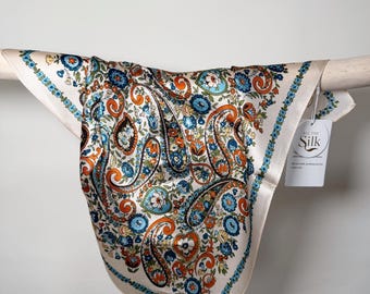 May include: A square silk scarf with a cream background and a paisley pattern in shades of orange, blue, and green. The scarf is draped over a wooden rod. A tag reads "All The Silk".