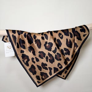Puede incluir: Un pañuelo cuadrado de seda con estampado de leopardo. El pañuelo presenta un fondo beige con manchas negras y marrones, y un borde negro. La etiqueta dice "All the Silk".