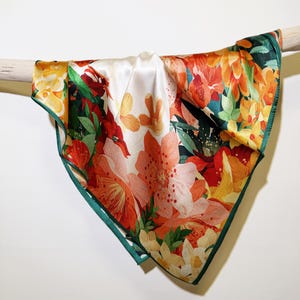 May include: A square silk scarf with a colourful floral design. The scarf displays large, stylised flowers in shades of orange, red, and yellow against a white backdrop. The edges are trimmed with a dark green border. The scarf is draped over a wooden rod.