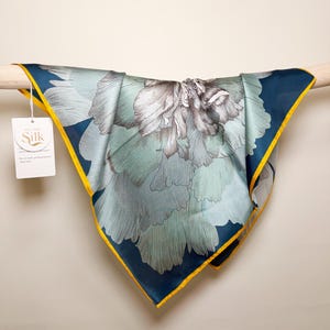 Pode incluir: Lenço de seda quadrado com um grande desenho floral em tons de azul, verde e cinza. O lenço tem uma borda azul marinho com uma borda amarela. Uma etiqueta branca com o texto "All the Silk" está anexada.