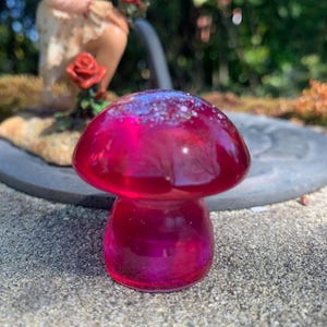 Puede incluir: Objeto decorativo en forma de hongo, de color magenta translúcido. El hongo tiene un sombrero redondeado con una parte superior moteada y brillante y un tallo. Se encuentra sobre una superficie texturizada, con un fondo borroso que incluye una estatua de hada y follaje. Mide unos 6 cm de alto.