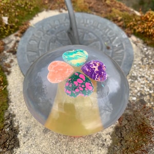 May include: A clear, spherical paperweight with four heart-shaped, colourful inclusions. The paperweight sits atop a stone surface, with a sundial in the background. The hearts are orange, green, purple, and teal.