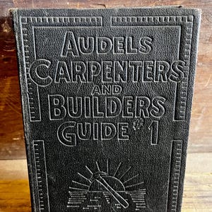 Pode incluir: Um livro preto intitulado "Audels Carpenters and Builders Guide #1". A capa apresenta texto em relevo e um gráfico de um martelo e sol. O livro é retangular e parece vintage.
