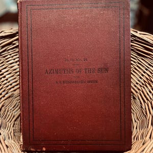 Op de afbeelding: Een vintage boek met een bordeauxrode kaft en zwarte rand, getiteld "Azimuths of the Sun". De tekst "U.S. HYDROGRAPHIC OFFICE" is ook zichtbaar. Het boek ligt in een gevlochten mand.