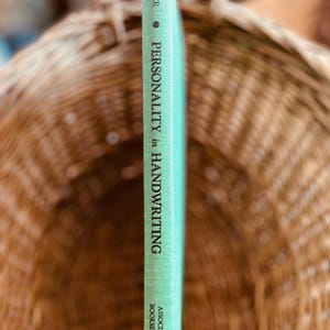 May include: A vintage book titled "Personality in Handwriting" by Singer. The book has a light green cover with black text. The book is set against a blurred background of a woven basket.