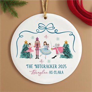 Może przedstawiać: Okrągła, biała ceramiczna ozdoba choinkowa z motywem Dziadka do Orzechów. Projekt zawiera ilustracje postaci baletowych, płatki śniegu oraz tekst "The Nutcracker 2025" i "Braylee as Clara". Do zawieszenia dołączona jest złota wstążka.