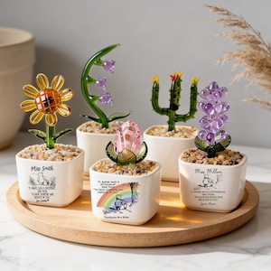 Puede incluir: Cinco figuras de cristal de flores y un cactus en macetas cuadradas blancas rellenas de pequeñas piedras. Las macetas presentan ilustraciones y citas de Winnie the Pooh. El conjunto incluye un girasol amarillo, campanillas moradas, una rosa rosa, un cactus verde y un jacinto morado, dispuestos sobre una bandeja redonda de madera.