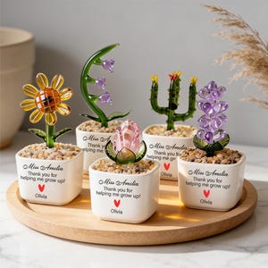 May include: Five decorative glass flower sculptures in small white pots filled with pebbles, arranged on a wooden tray. Each pot has the text "Miss Amelia, Thank you for helping me grow up!" and the name "Olivia". The flowers include a sunflower, a rose, and a cactus.